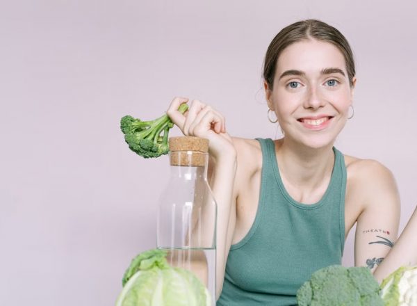Woman in tank top smiling with broccoli, cabbage, and glass of water on table.